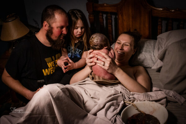 Mom is laying in bed, holding baby up in front of her to discover if it is a boy or girl.  She has a big smile on her face.  Dad and big sister are to the side of the bed.  Dad has a big smile, and big sister looks slightly confused or concerned. The placenta is still attached to baby and sits in a bowl next to Mom. 