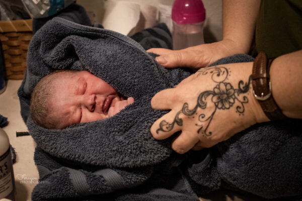 Midwife with tattoo on her hand is wrapping newborn in a blue towel after her first bath. 