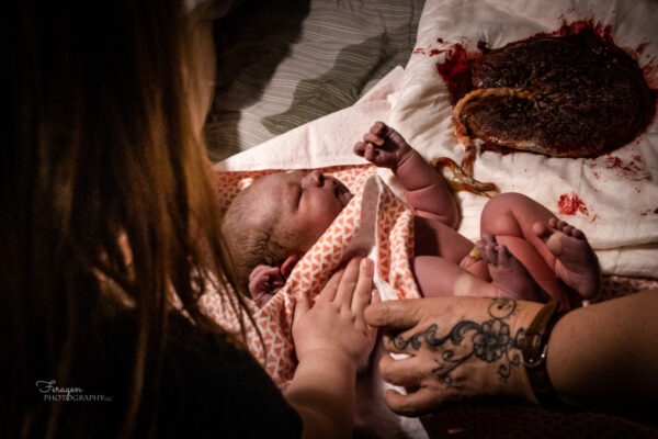 Newborn baby laying on an orange and white blanket on parent's bed.  Big sister and Midwife with tattooed hand are in foreground drying baby off.  The attached placenta and umbilical cord can be seen next to the newborn.