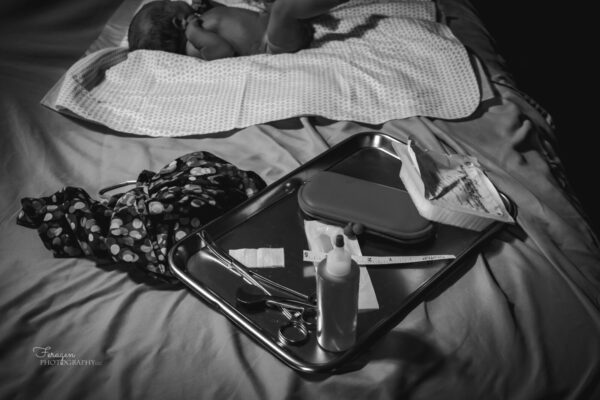 Black and White Photograph of Midwife equipment on a tray on the bed with weighing sling next to it.  Newborn can be seen in the background laying on patterned blanket.