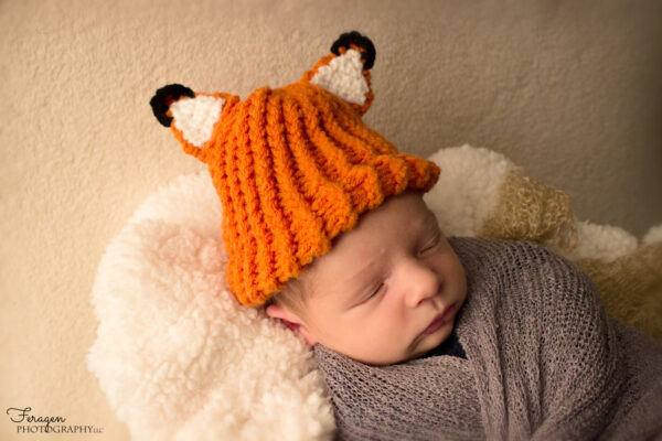 Newborn baby wearing a fox knitted hat is drifting off to sleep.