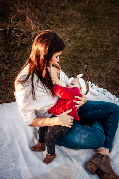 Mother in white sweater and jeans sitting on a white blanket breastfeeding her baby.