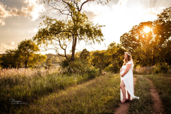 Pregnant woman in white maternity gown holding her stomach while looking into the distance with trees, sun, and lake in the background.
