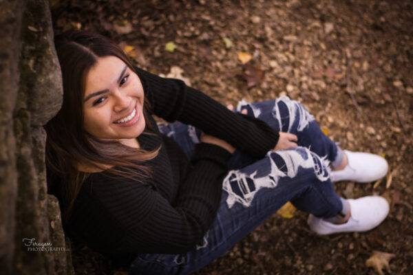 High School Senior sitting on leaf covered ground smiles up at camera.