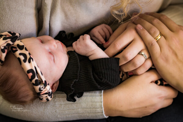 Newborn in cheetah print headband is held by her mother.  Dad's hand with wedding ring is on top of Mom's hand with wedding right, and both hands are gently on top of baby. 