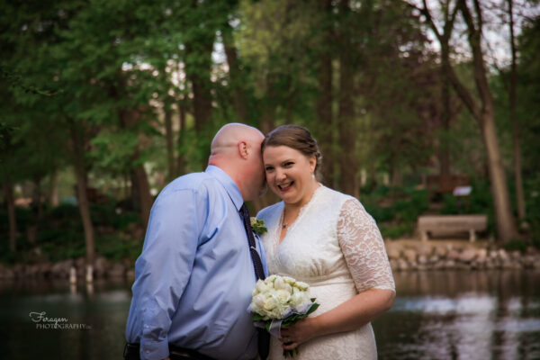 Bride laughs as new husband tells her a secret in front of a lake.